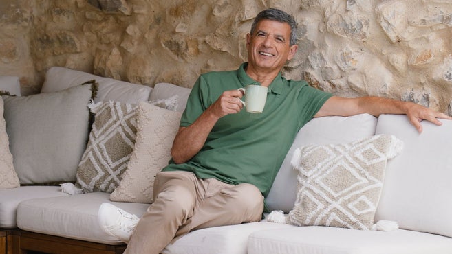 Um homem sorridente sentado confortavelmente num sofá de exterior com almofadas brancas, a segurar numa caneca de cerâmica. Está a usar um polo verde e calças beges, inclinando-se contra um fundo de parede de pedra.