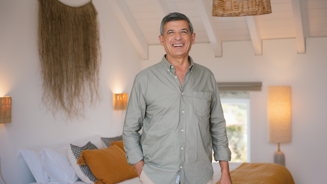 Um homem sorridente e confiante a usar uma camisa verde, num quarto moderno e bem iluminado com iluminação quente, uma parede tecida pendurada e roupa de cama amarela mostarda.