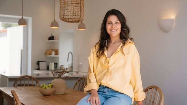 Uma jovem sorridente com uma camisa amarela e calças de ganga azuis sentada numa mesa de jantar de madeira numa cozinha e sala de jantar modernas e abertas.