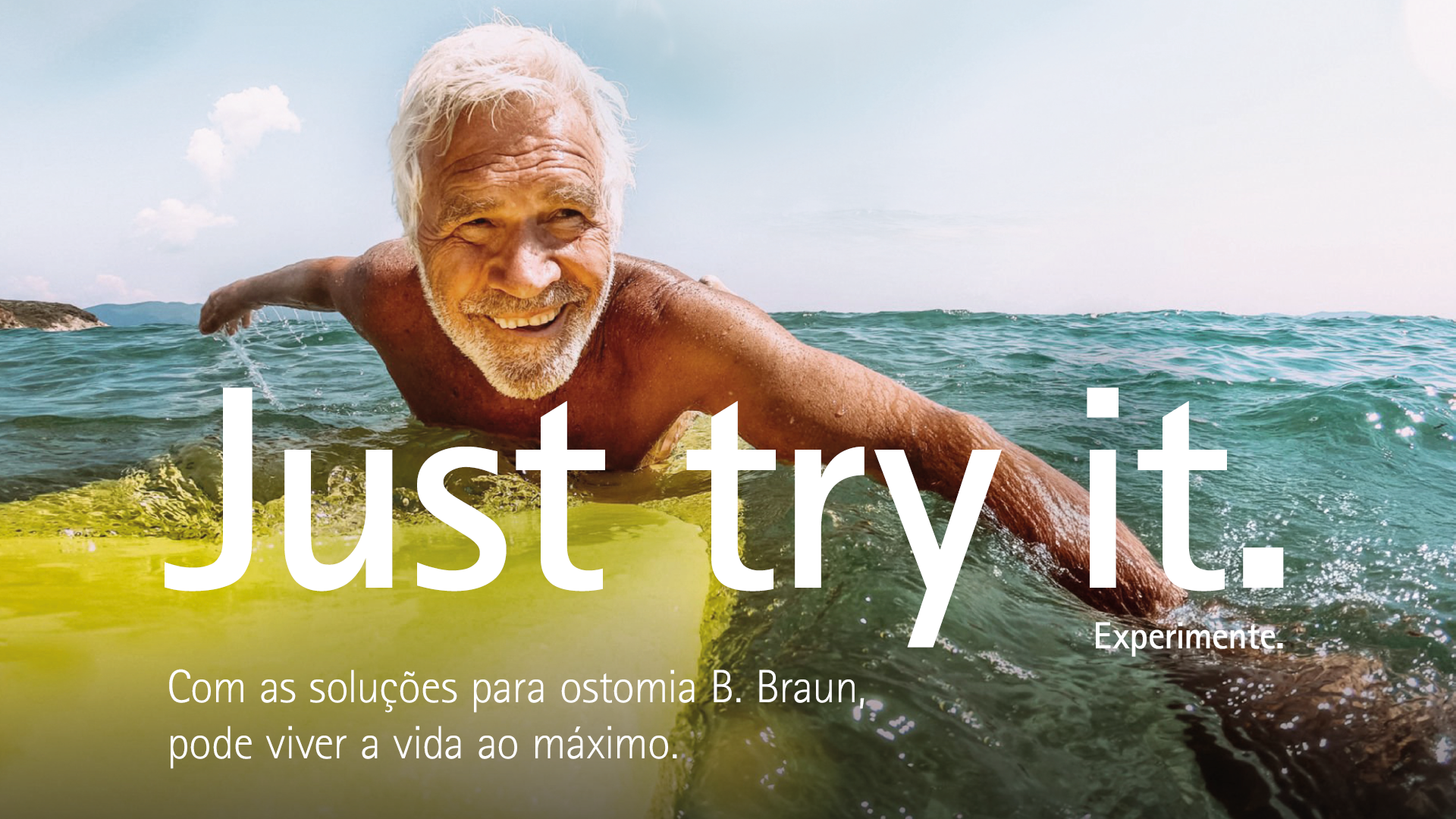 An elderly man with white hair is lying on a yellow surfboard, paddling in the ocean under a clear blue sky.