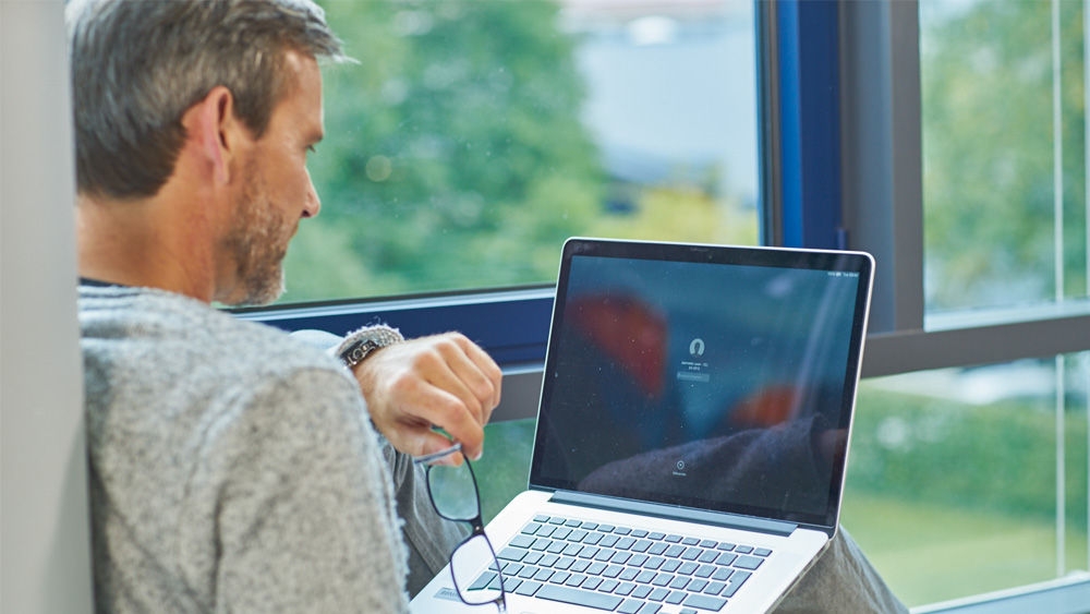 Uma pessoa sentada segura um computador portátil ao colo. Com a mão esquerda, estão a escrever no teclado, enquanto seguram um par de óculos na mão direita. No ecrã do computador portátil, é visível um símbolo desfocado que se assemelha a auscultadores, juntamente com a etiqueta "Toque para ouvir". Em segundo plano, é possível ver uma paisagem verdejante e natural através de uma janela grande.