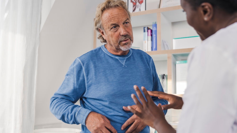 um homem de meia idade faz uma pergunta ao médico enquanto segura o lado direito do estômago, um médico explica-lhe algo