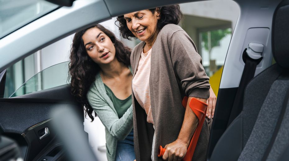 Um Doente que usa uma muleta e é ajudado por outra pessoa a entrar no seu veículo.