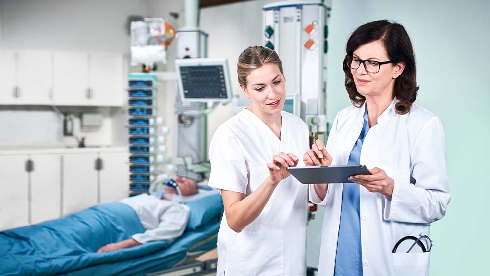 Um médico e um enfermeiro estão a discutir a dosagem para a infusão inteligente da B. Braun, que está a ser introduzida diretamente a partir de um comprimido. O Doente na UCI pode ser visto em segundo plano, ligado a uma torre cheia de dispositivos de infusão inteligentes.