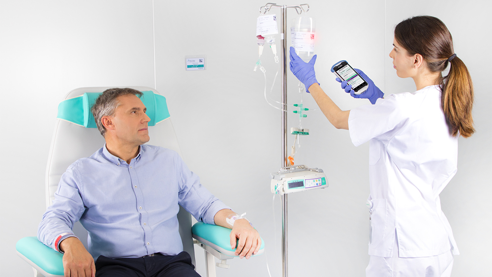 A imagem mostra um Doente sentado numa cadeira, a receber uma infusão de um dispositivo médico da B. Braun, enquanto um enfermeiro está a configurar a dosagem de infusão no dispositivo.