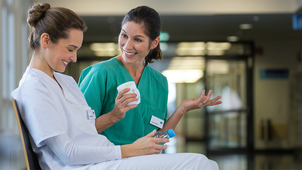 Duas enfermeiras sentadas no corredor do hospital a fazer uma pausa para beber café e água. Ambos sorriem enquanto falam.  