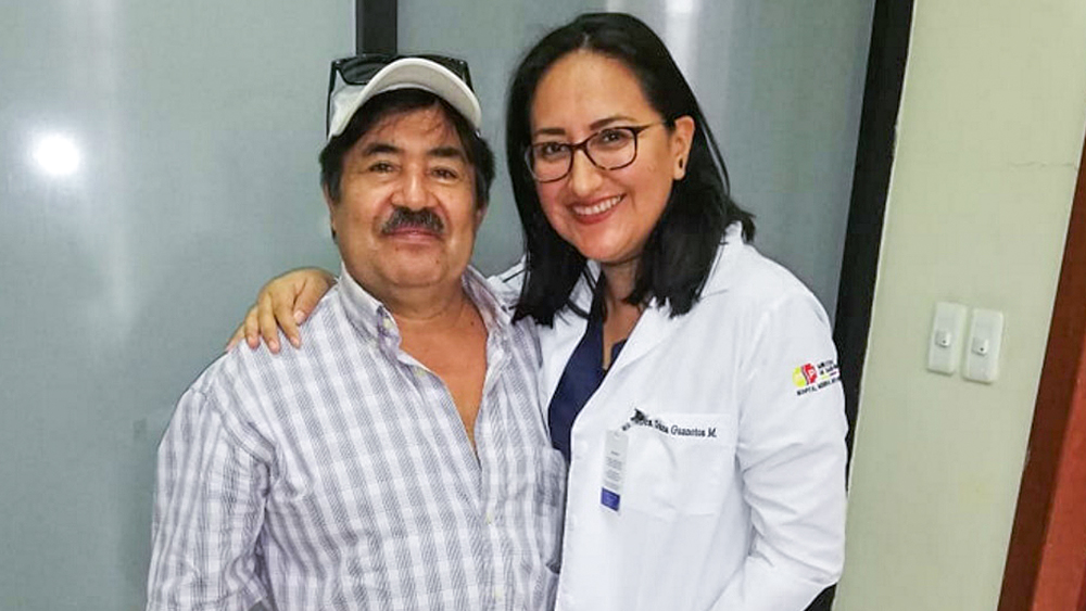 Um homem e uma mulher estão um ao lado do outro e sorriem. A mulher segura o braço à volta do homem