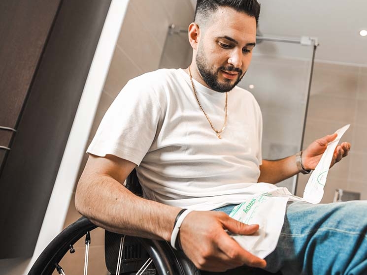 Um homem numa cadeira de rodas a segurar um cateter na mão, e uma casa de banho em segundo plano