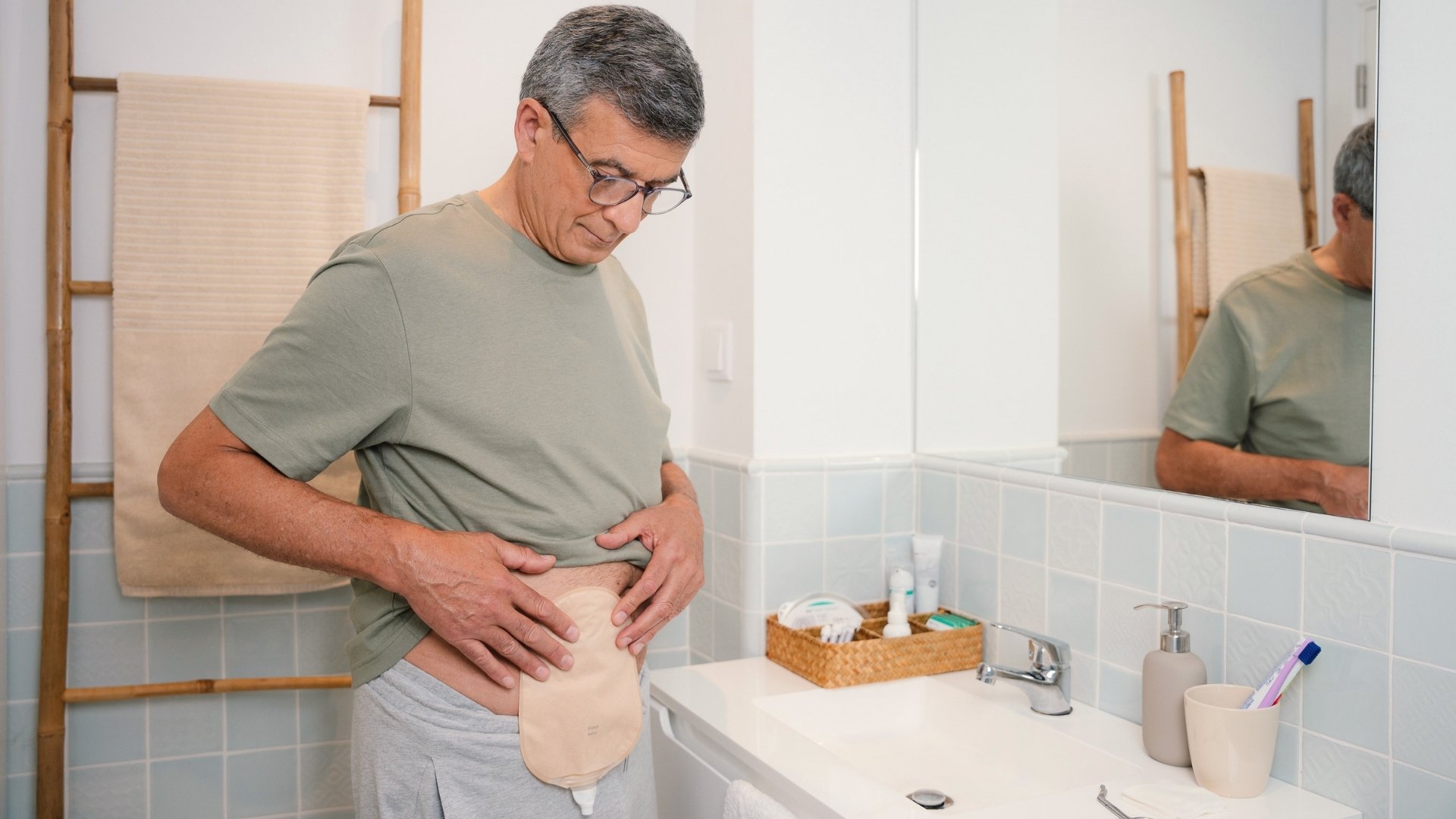 Um homem de óculos e t-shirt verde olhando para baixo para a sua bolsa de urostomia presa ao abdómen enquanto está de pé numa casa de banho. Ele tem o seu reflexo visível num espelho ao seu lado.