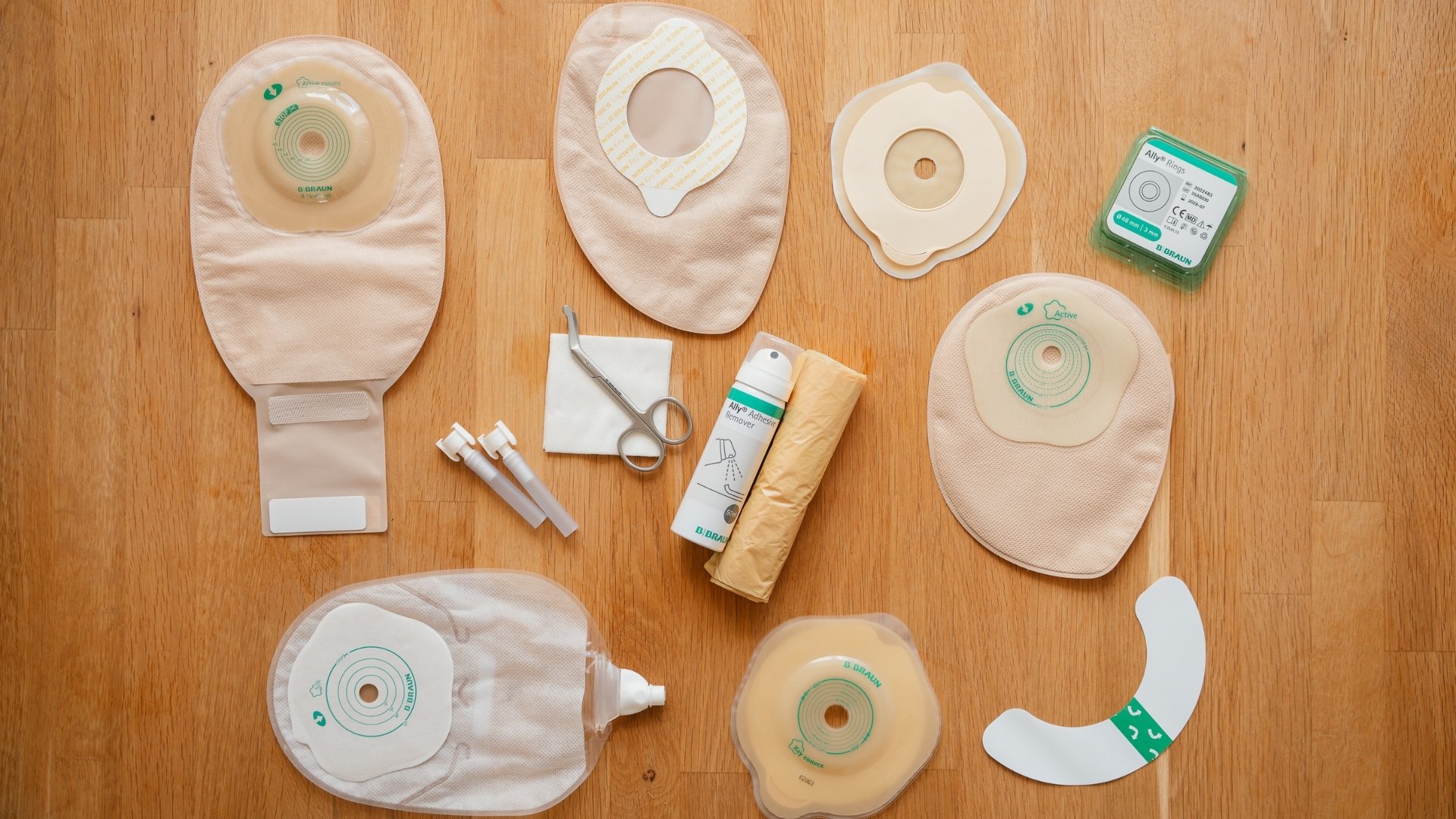 Vista aérea de produtos e acessórios para estoma da B. Braun dispostos sobre uma superfície de madeira clara. A coleção inclui diferentes tipos de bolsas bege para estoma, placas para estoma, barreiras cutâneas, anéis para estoma, tesouras médicas curvas, spray removedor de adesivo e saco para descarte.