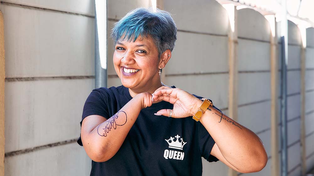 Uma mulher sem mão direita, cabelo azul e tatuagens sorri, irradia confiança e auto-expressão. 